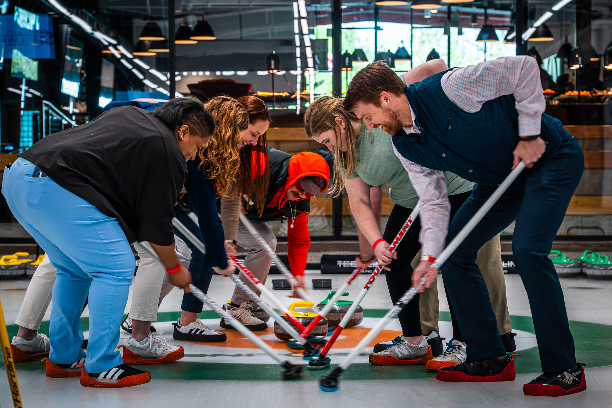 INSBANK Group of people bending over with floor hockey sticks in a circle indoors, focused on the puck/ball in the center.