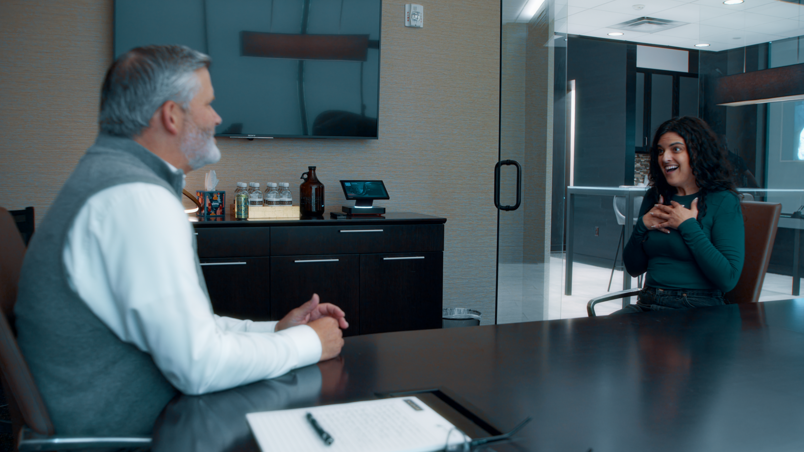 INSBANK Two people sit at a dark conference table in a modern office, talking; man with gray hair on the left, smiling woman on the right.
